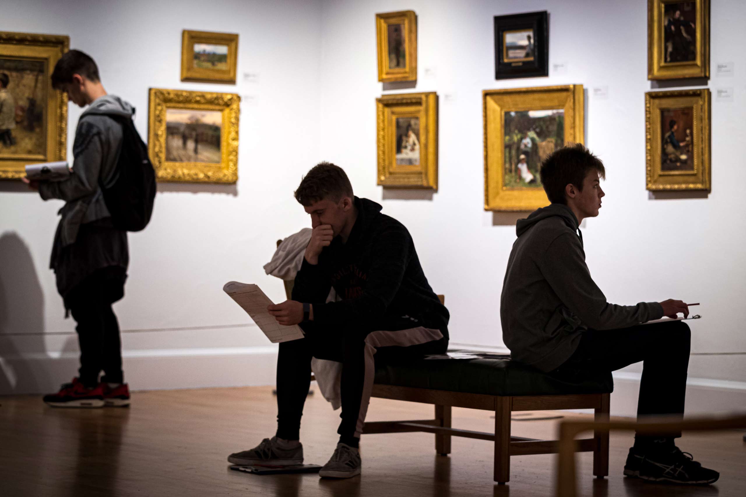 Two schoolchildren sit on a bench in the museum while another student in the background studies a painting on the wall