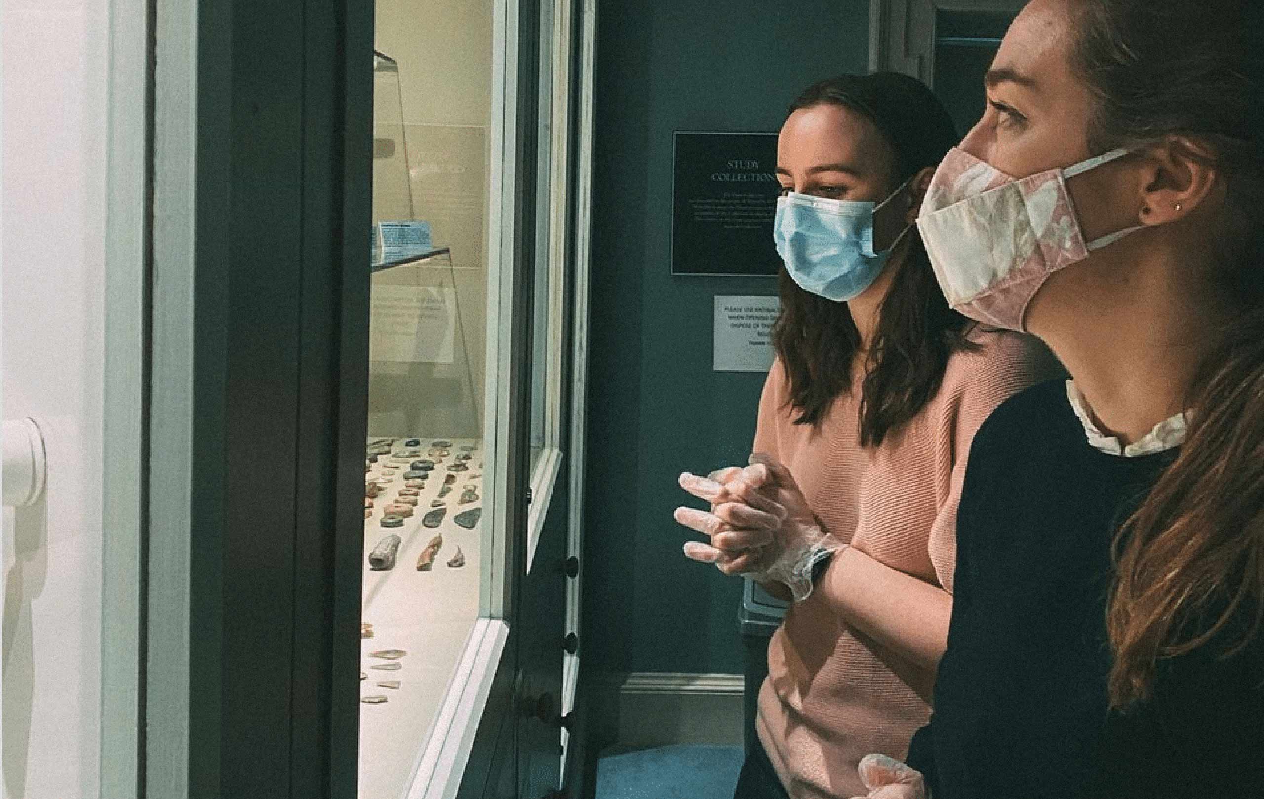 Two girls wearing masks and disposable gloves looking at a glass exhibition