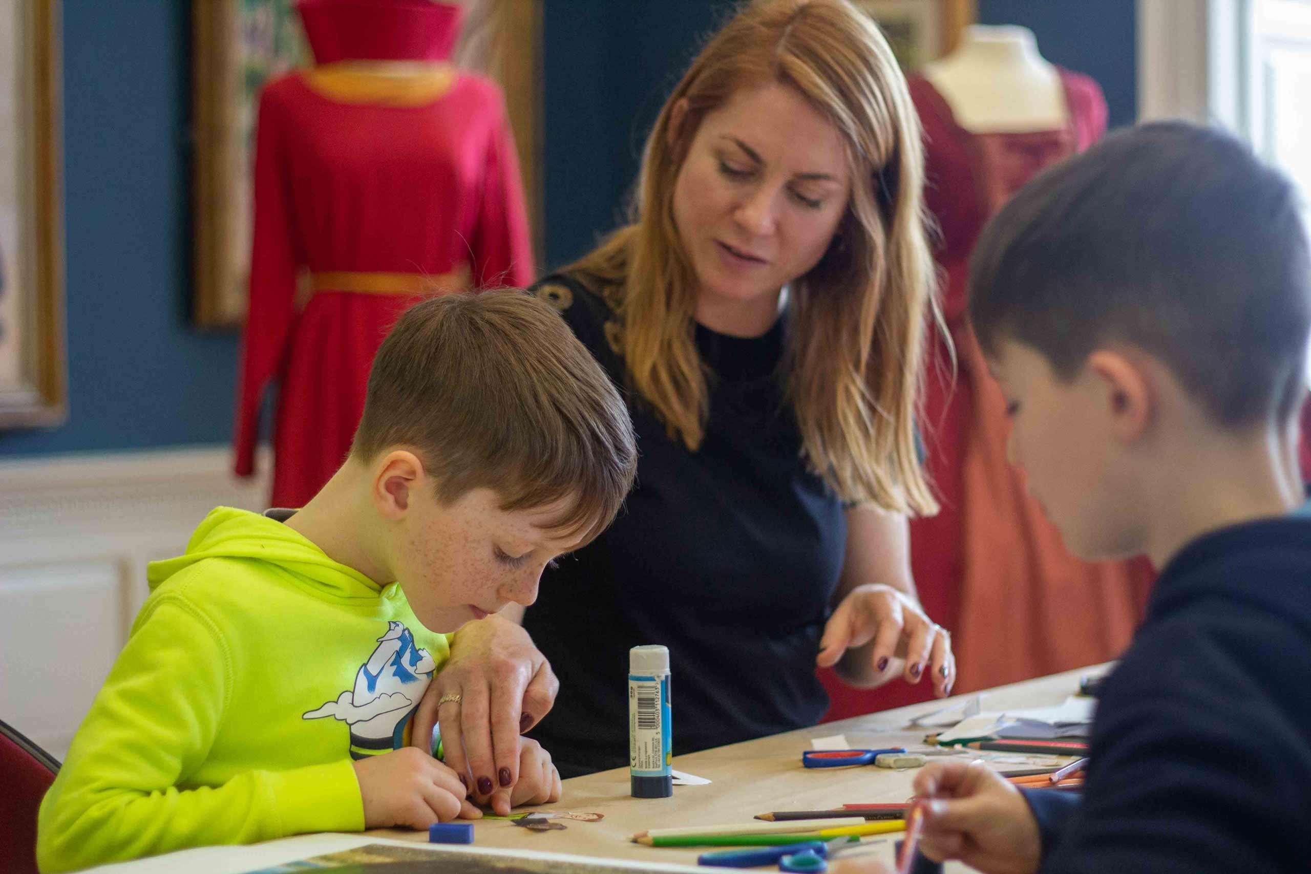 A woman sits with two bots at a table with art supplies and helps them glue some cutout paper together