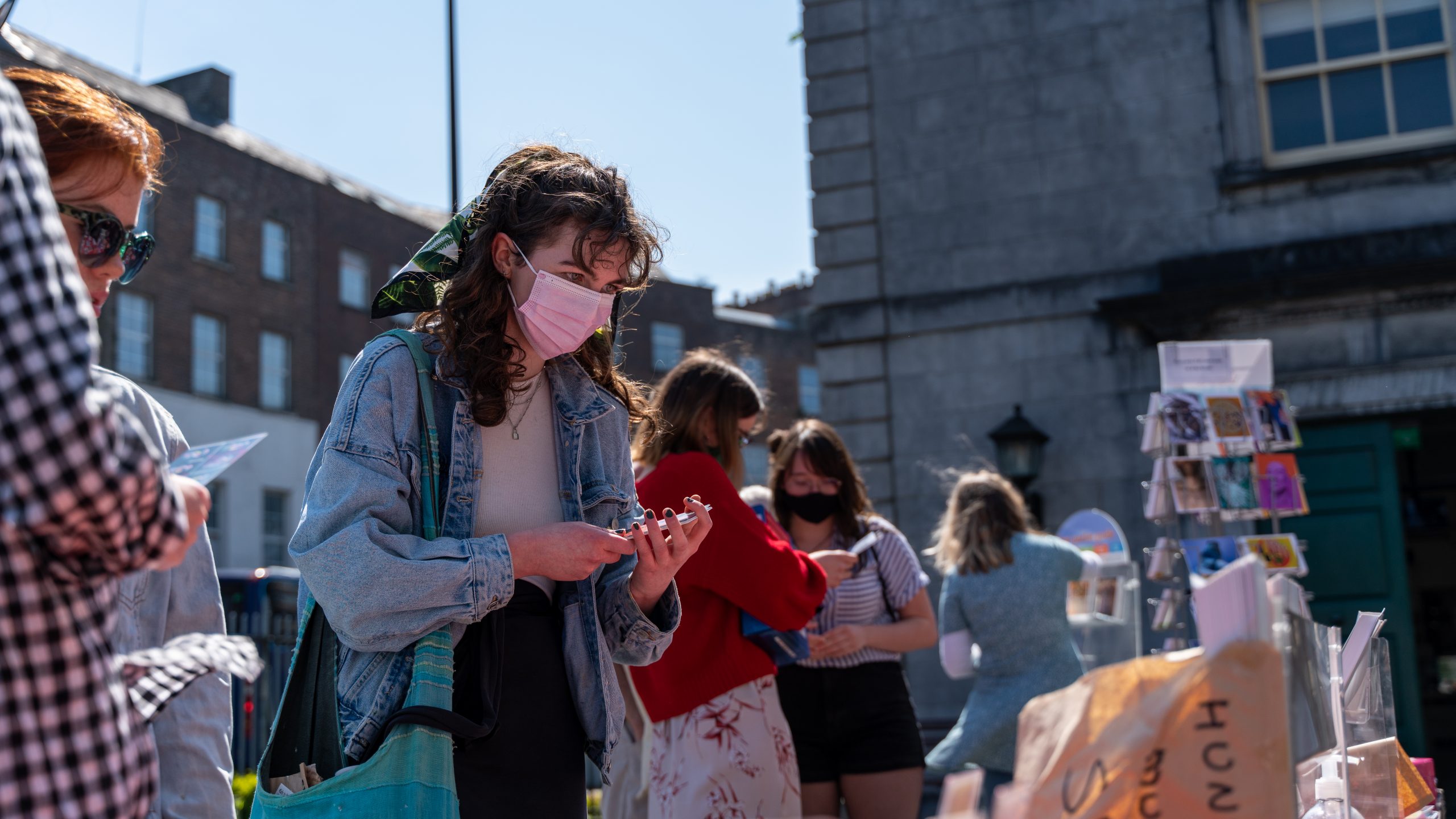 A girl in a denim jacket and a pink disposable mask holds a stack of postcards
