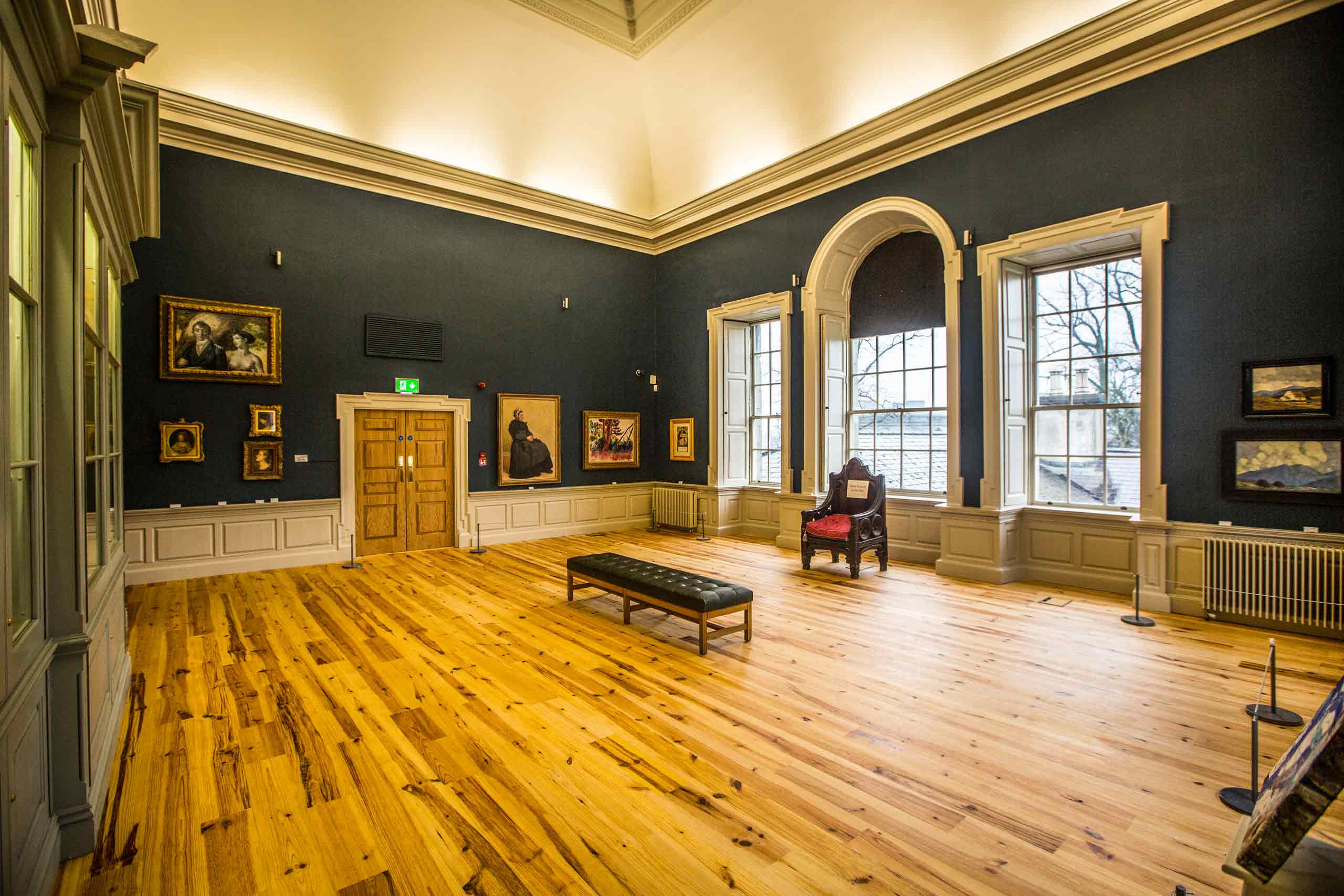A large room with paintings on the wall in the Hunt Museum. There is a bench in the middle of the room and a chair in front of 3 large windows.