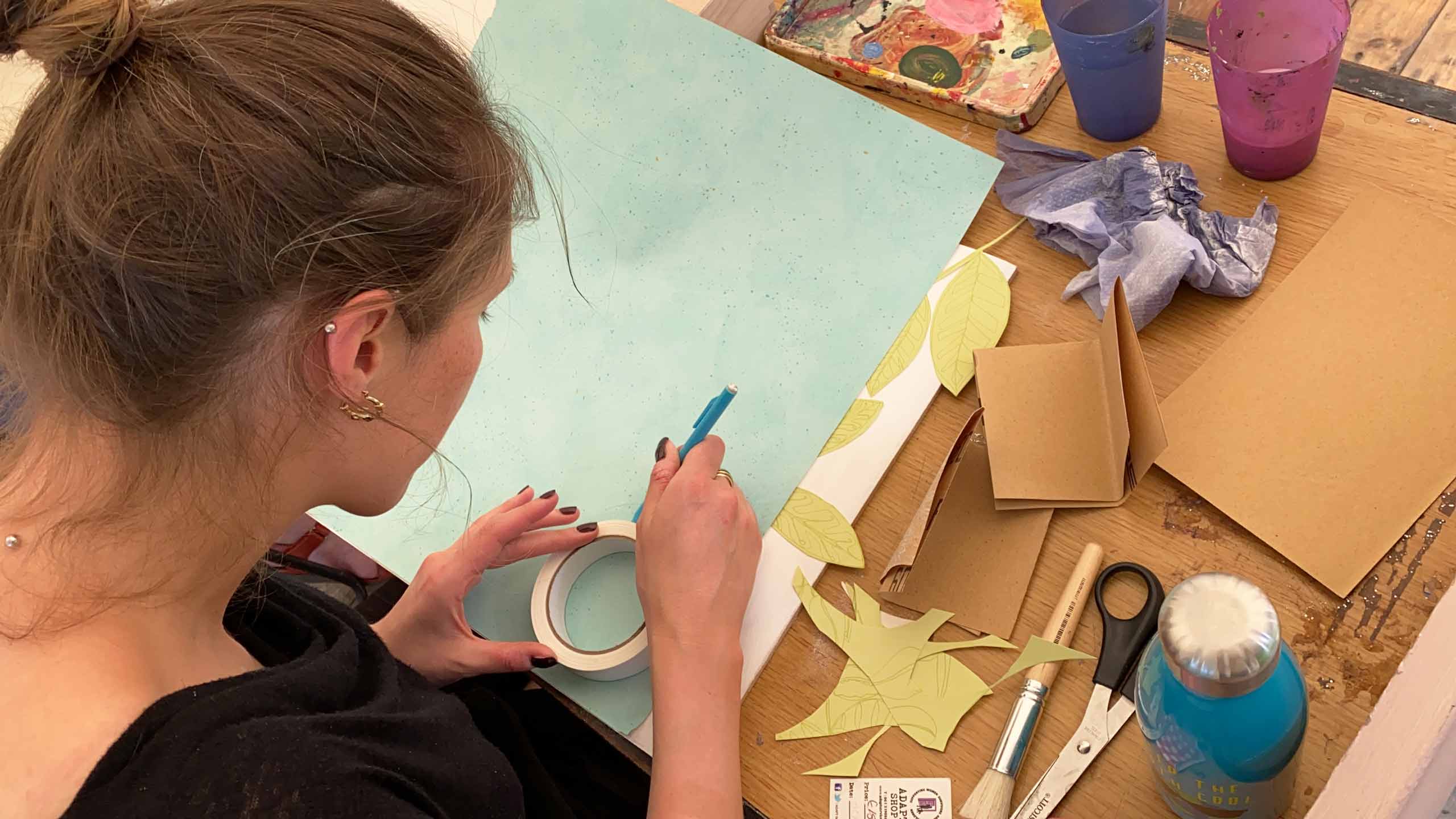 A woman traces around a roll of masking tape with pencil onto a piece of blue card. Around her on the table are various art supplies.Museum from Home