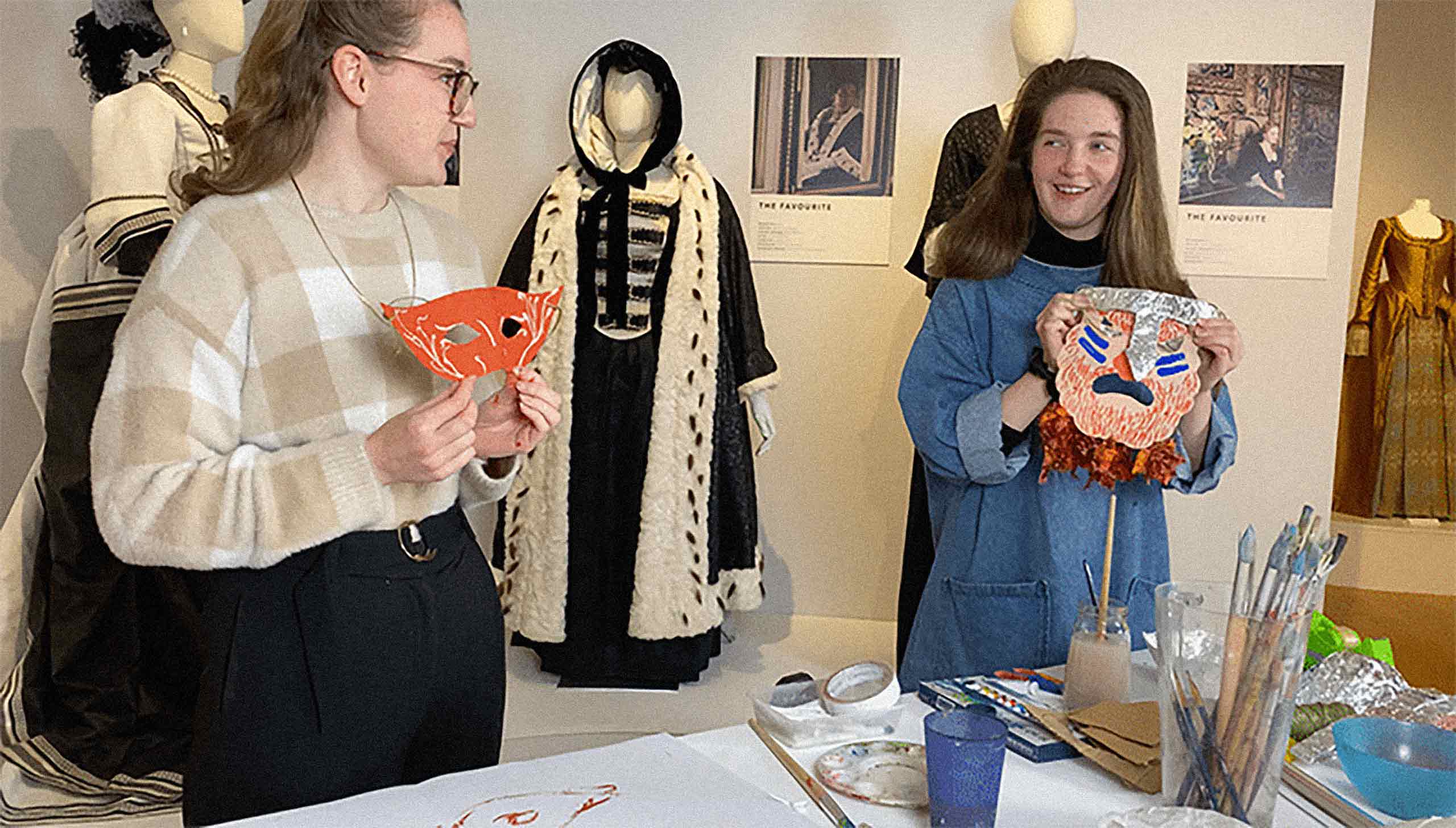 Mask . Two teenagers stand at an art and crafts table and hold up two handmade masks. Behind them are mannequins wearing pieces from different time eras.