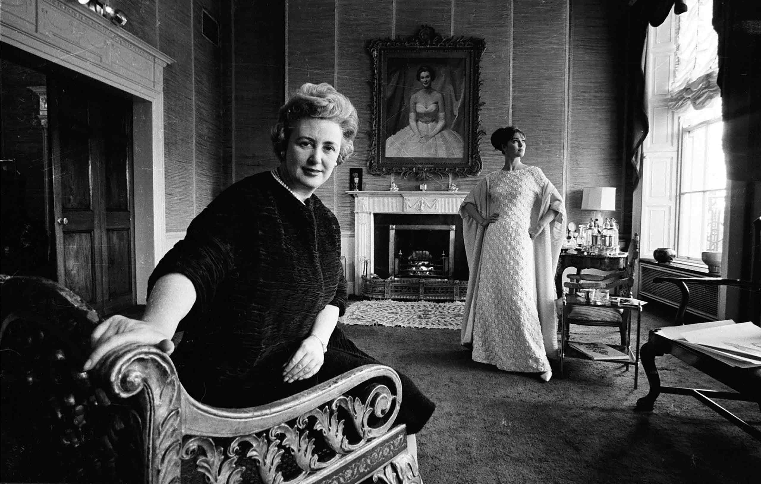 Sybil Connoly in an armchair in a grand room with a large window and fireplace. Above the mantlepiece is a portrait of a woman in a white dress, and in front of the mantlepiece a woman poses looking out the window in a white dress.